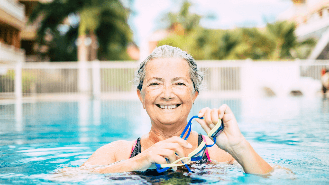 A woman with arthritis standing in a pool, wearing a black and pink swimsuit and holding swim goggles. She is looking directly at the camera and smiling.